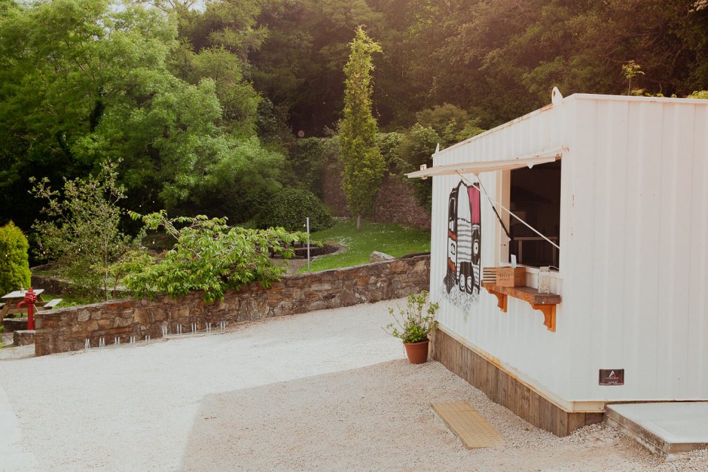 Cream shipping container/ kitchen with a picture of a horsebox on the side. Trees in background and white stones on ground.